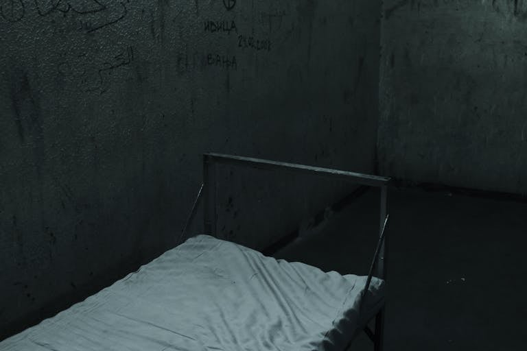 Metal Bed in a Concrete Prison Cell with Names Scribbled on Walls