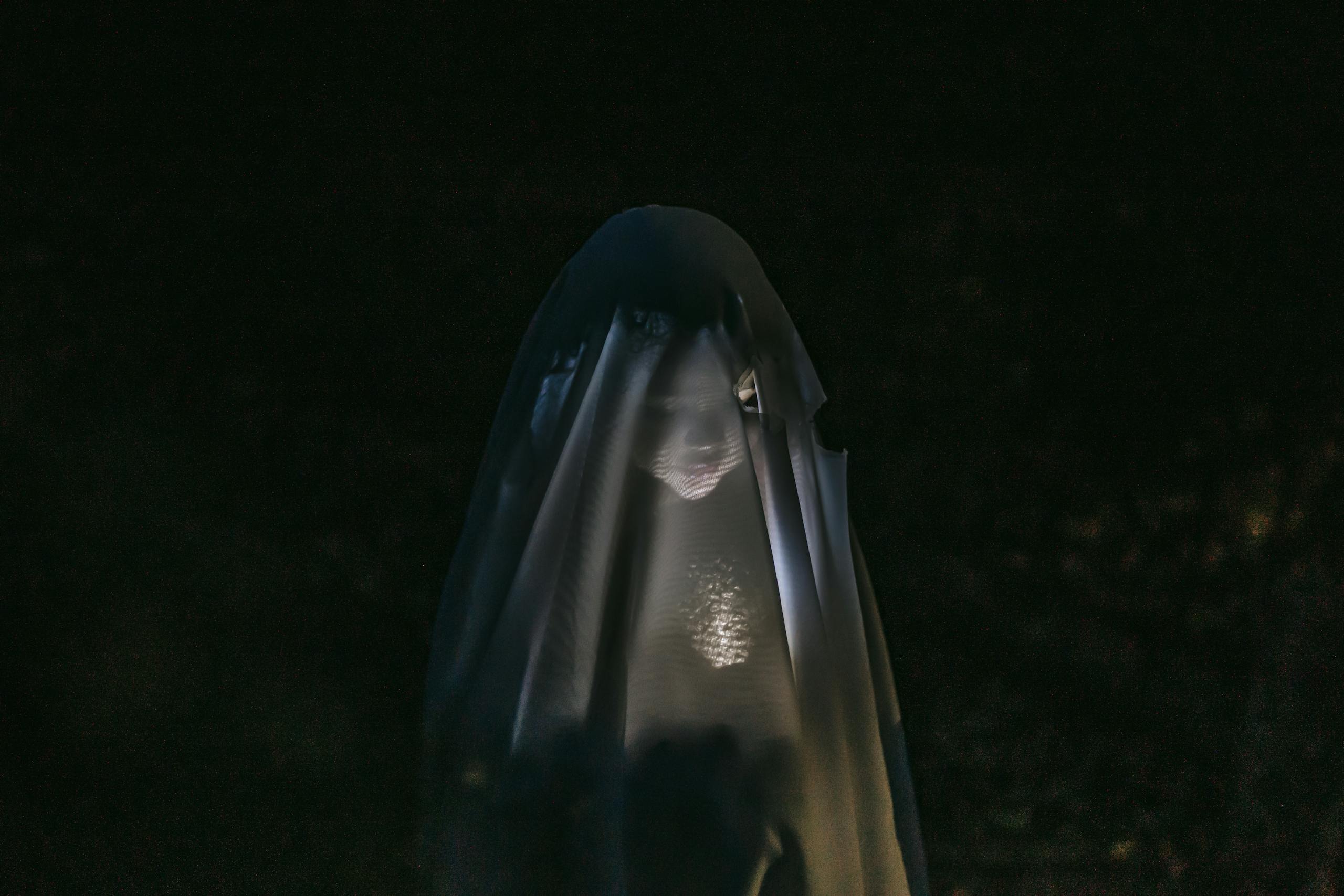 Mysterious little girl standing in darkness covered with white blanket as ghost and shining flashlight on face
