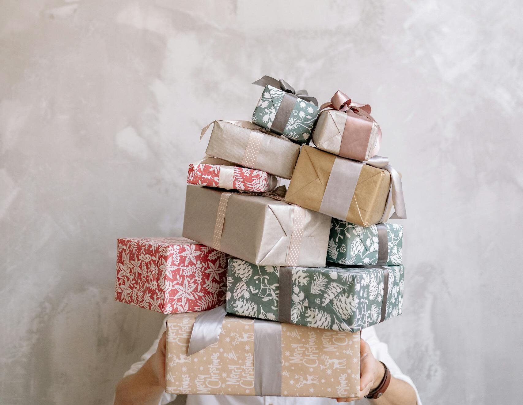 A person holding a stack of beautifully wrapped gifts against a textured wall, showing festive celebration.
