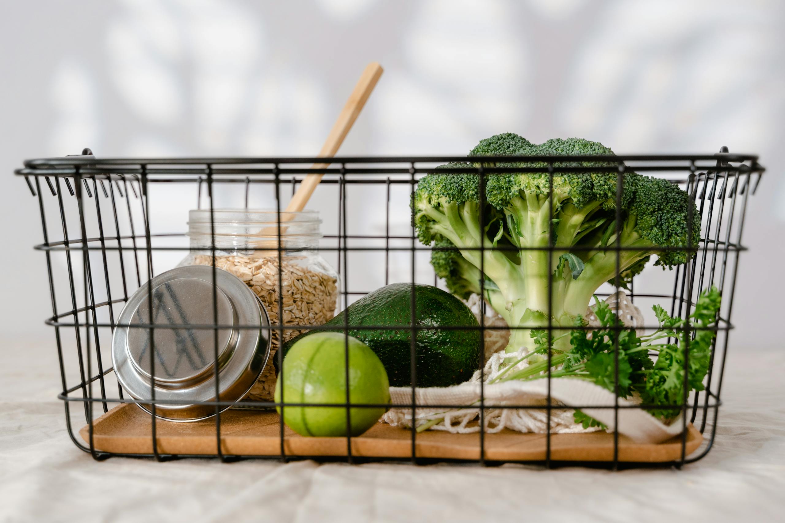 A wire basket holds fresh broccoli, lime, avocado, oats in a jar, and leafy greens.