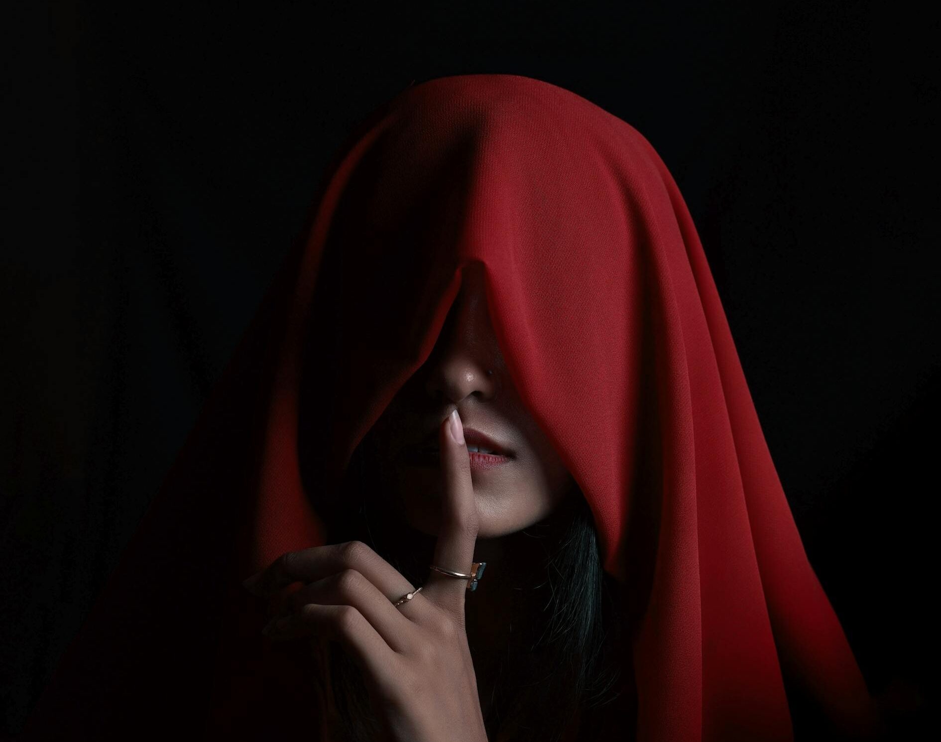 Mysterious woman in a red hood, finger on lips for silence, against a dark background.